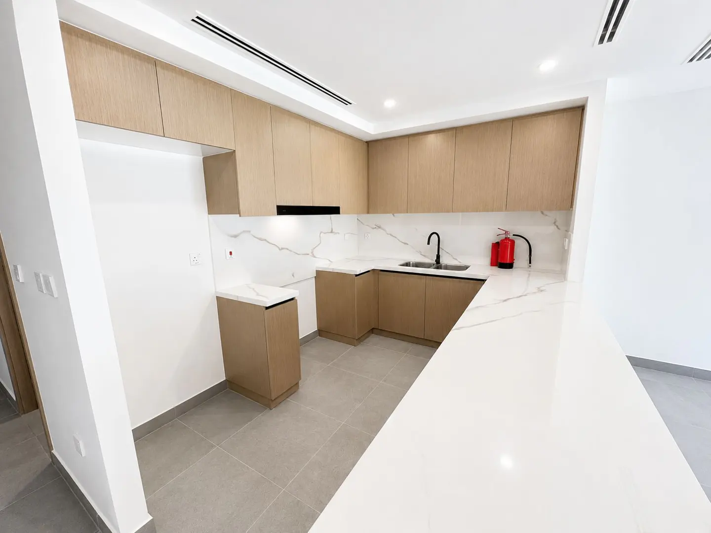 Bright kitchen with light wood cabinets, white marble countertops and backsplash, black faucet, and a red fire extinguisher.