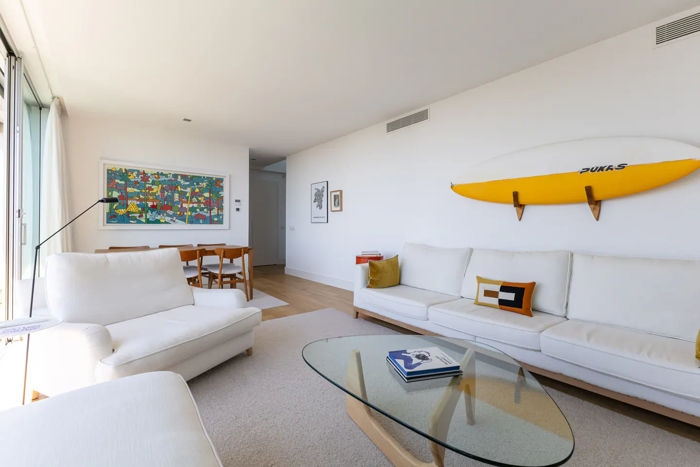Bright living room with white sofa, glass table, and yellow surfboard on the wall. Dining table and colorful art in the background.