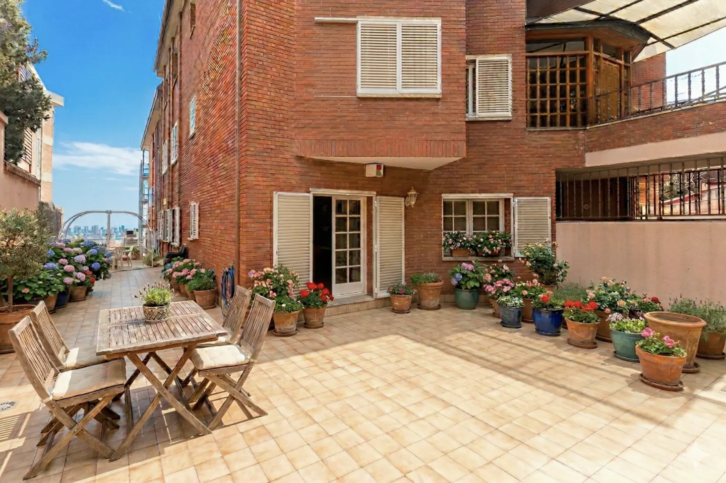 Brick building with a patio featuring a wooden table and chairs, potted flowers, and a view of the city skyline in the background.