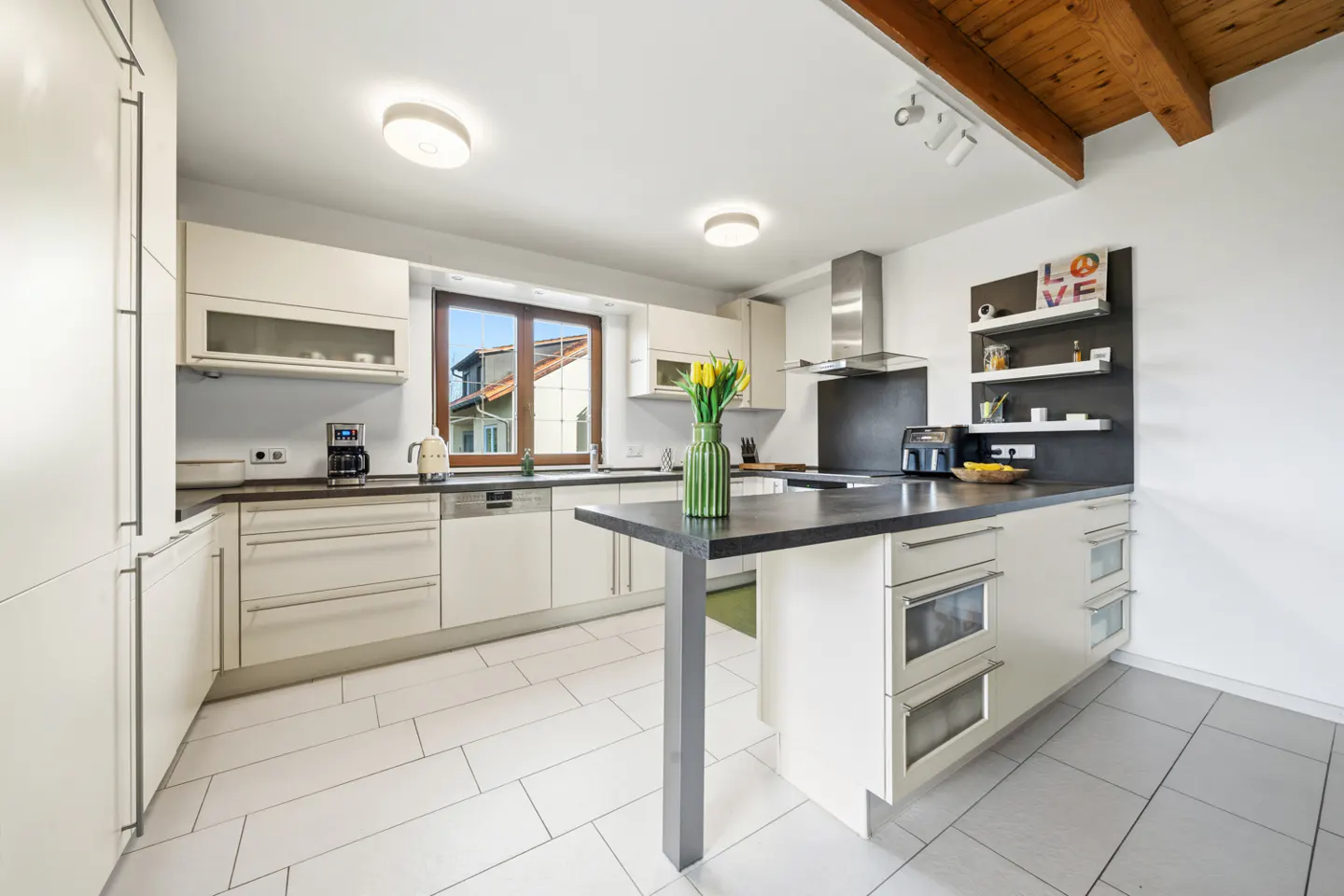 Bright kitchen with white cabinets, dark countertops, and a central island. A window overlooks a house. Yellow tulips add a pop of color.