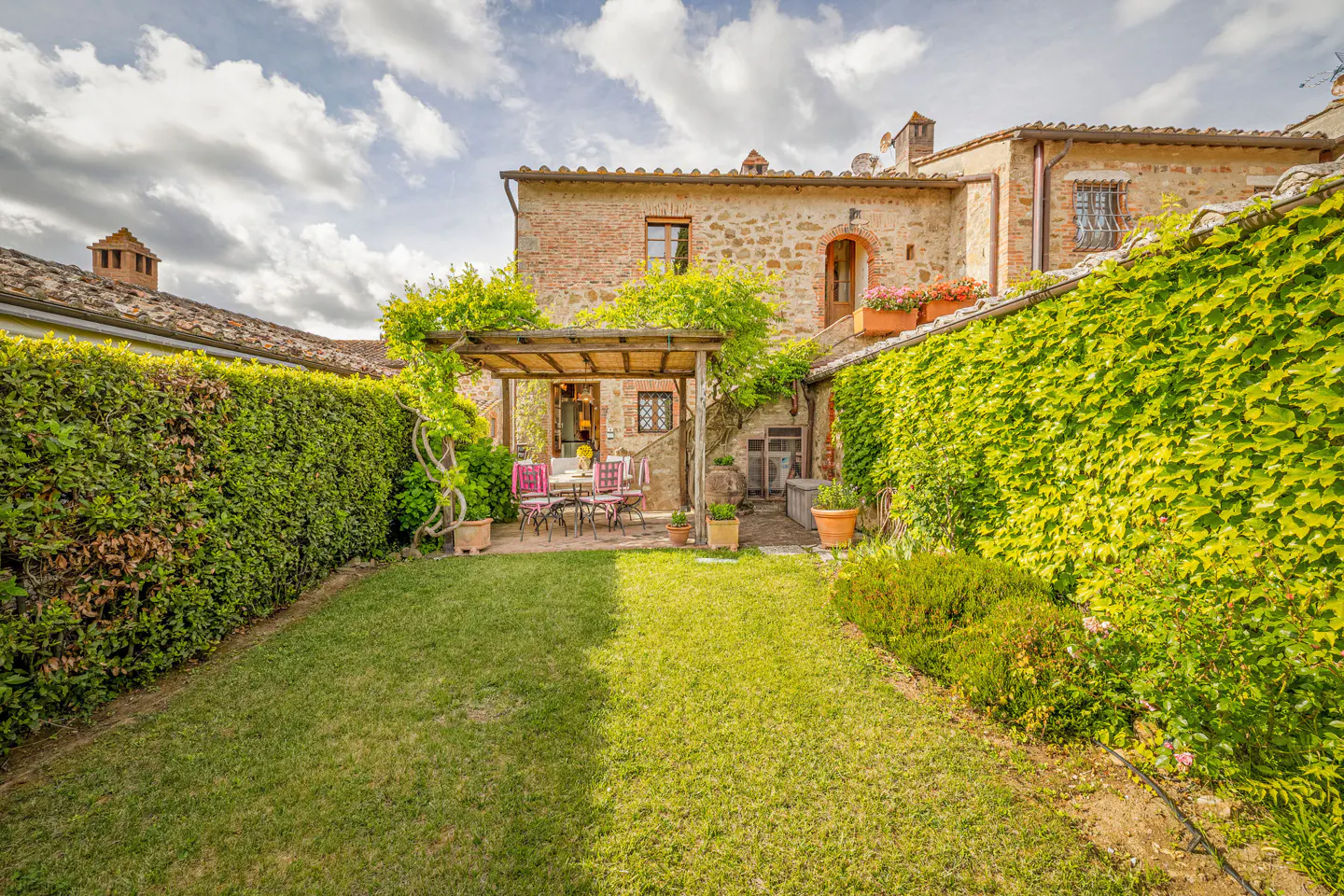 A stone villa courtyard with a green lawn, ivy-covered walls, and a wooden pergola over a patio dining set.