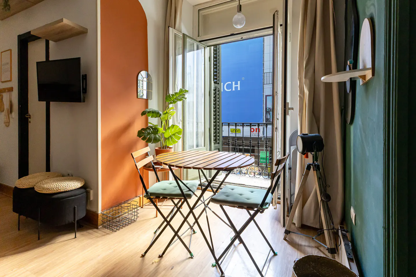 Cozy apartment interior with a round wooden table, two chairs, and an open balcony overlooking a city street. The walls are painted in orange and green.