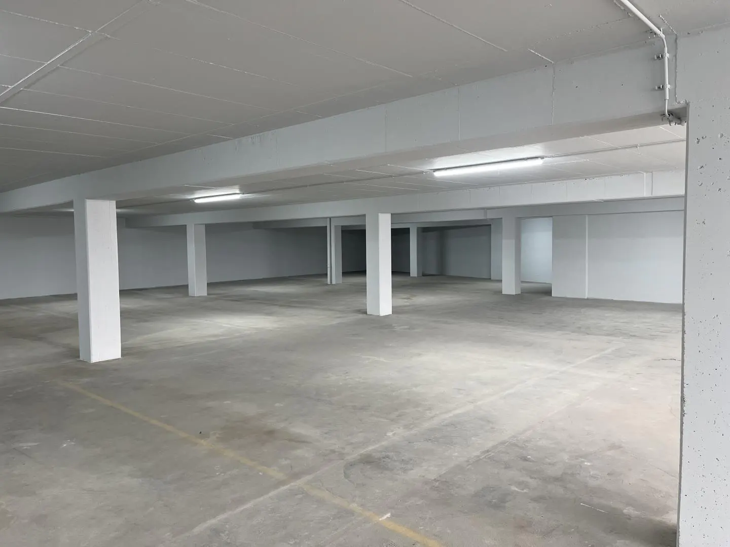 Empty parking garage with concrete floor, white walls and support columns. Overhead fluorescent lighting.