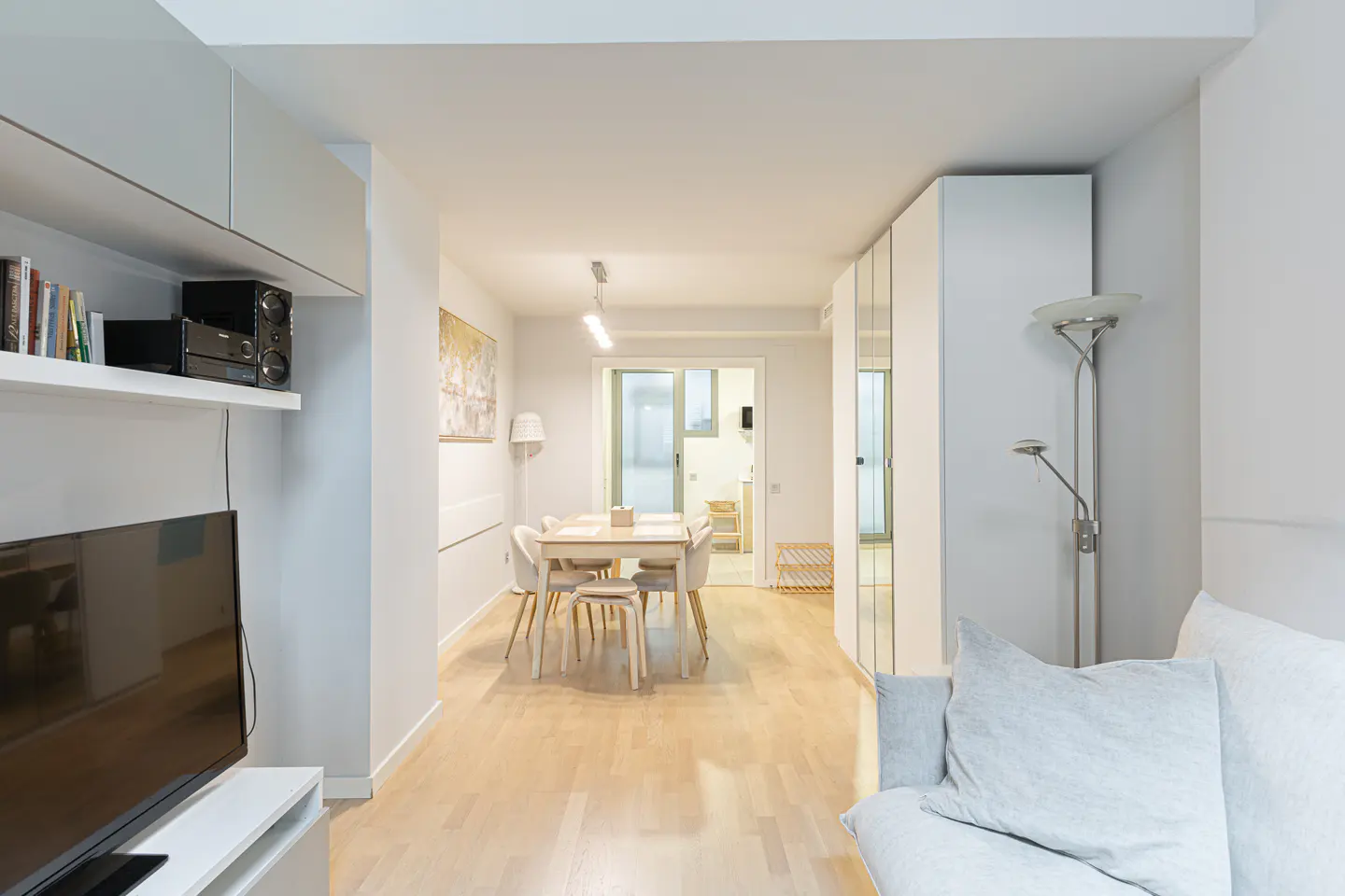 A bright, modern apartment interior with a dining table, TV, and light wood floors.