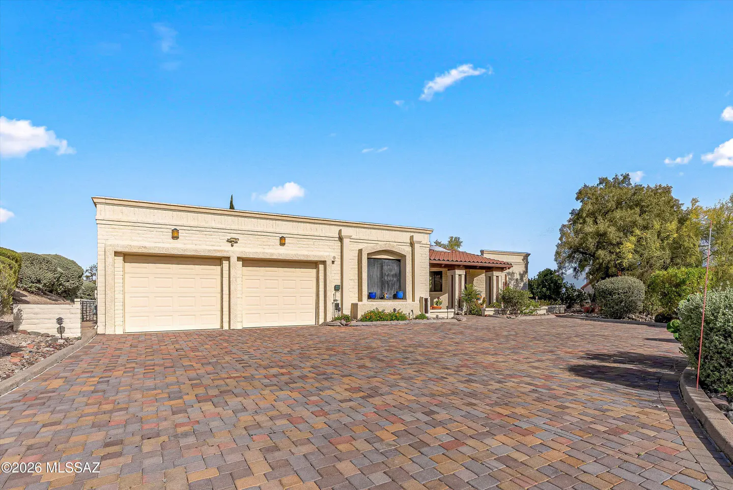 Beige brick house with a red tile roof and a two-car garage, viewed from a brick driveway under a blue sky.