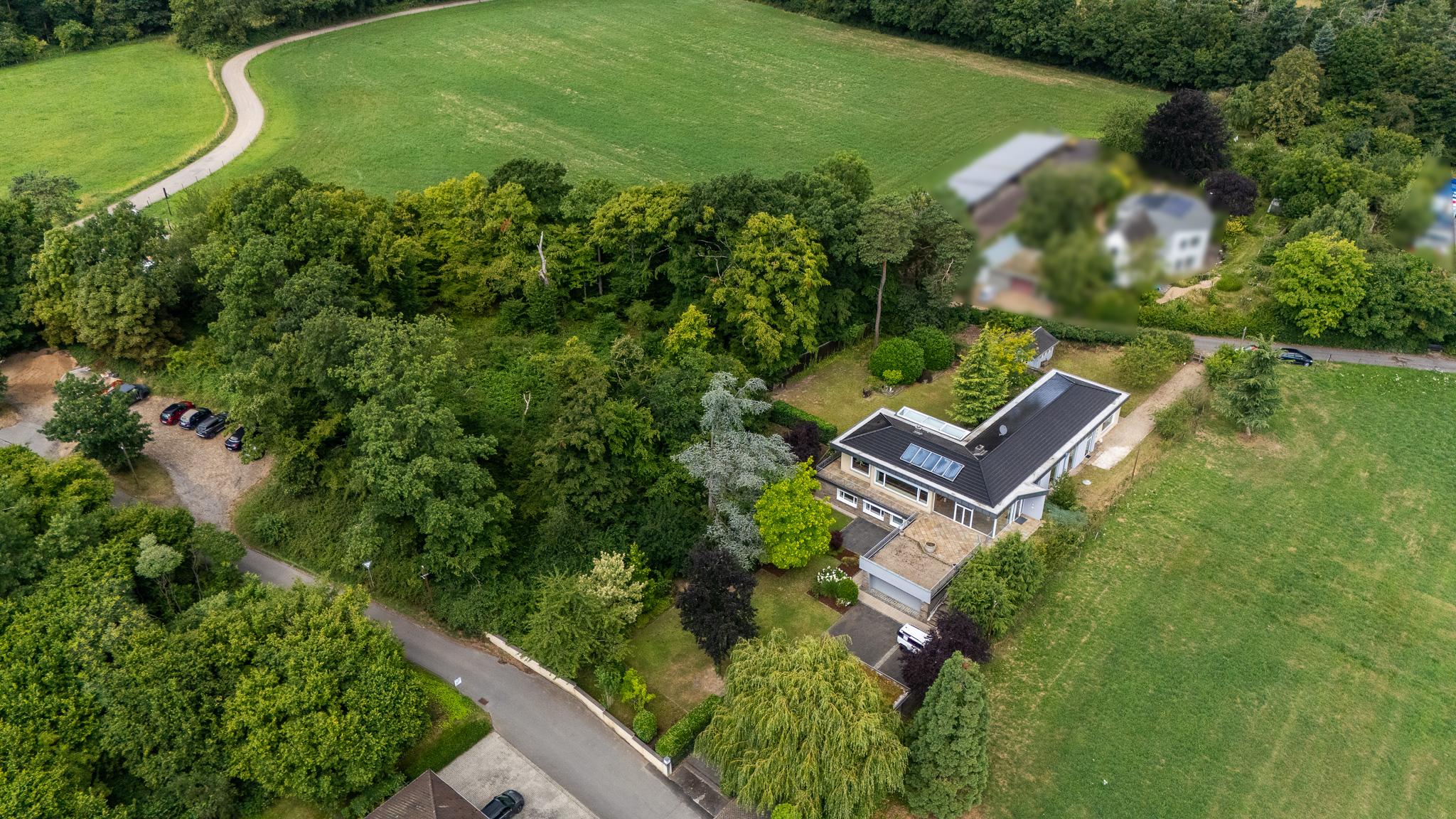 Aerial view of a modern house with a dark roof nestled in a lush, wooded area surrounded by expansive green fields.