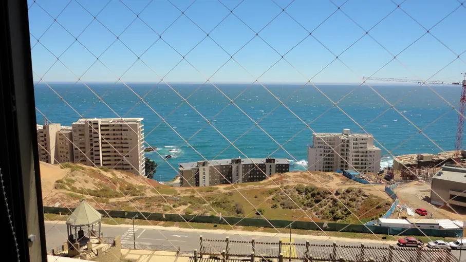 View from a window with a safety net, overlooking buildings, a road, and the ocean under a clear blue sky. A construction crane is visible in the distance.