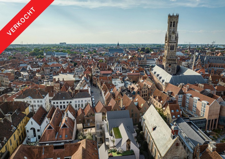 Aerial view of Bruges, Belgium, featuring red-tiled rooftops, a tall stone tower, and a red "Verkocht" banner.
