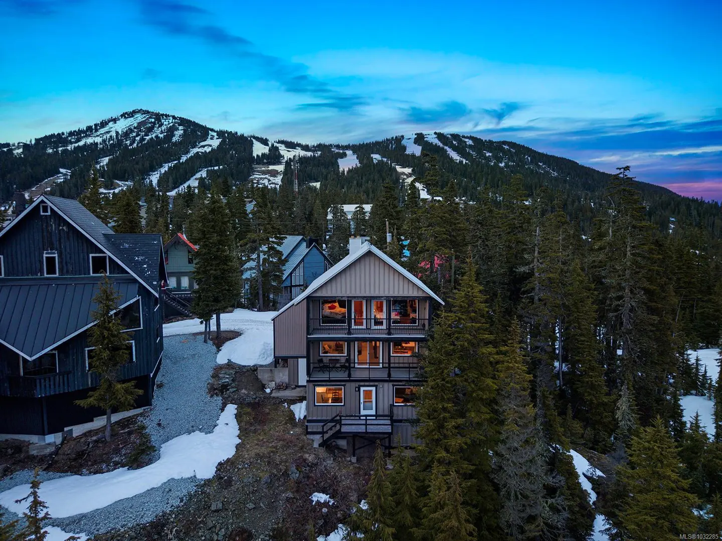 Three-story mountain home with balconies, nestled in trees with a snowy mountain backdrop under a blue sky.