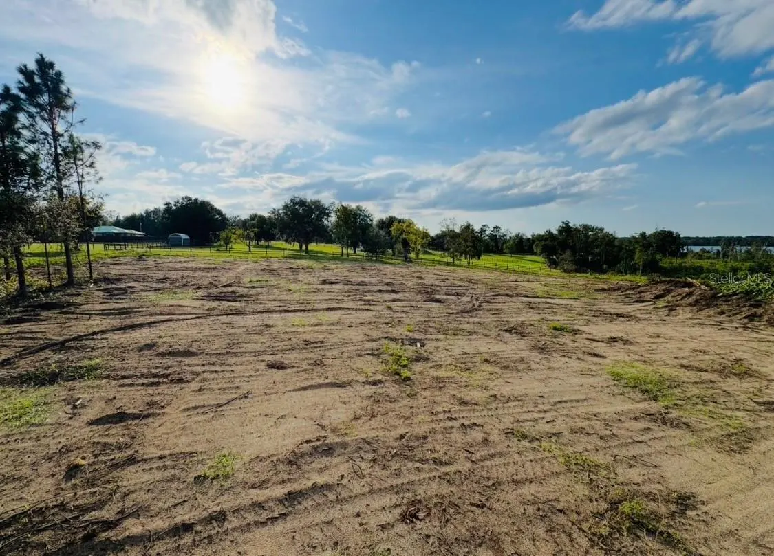 Vacant lot with tire tracks under a blue sky with scattered clouds. Trees line the back of the property.