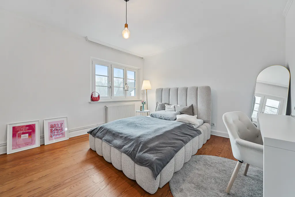 Bedroom with a gray bed, white walls, and wood floors. A desk and mirror are on the right, and framed art is on the left.