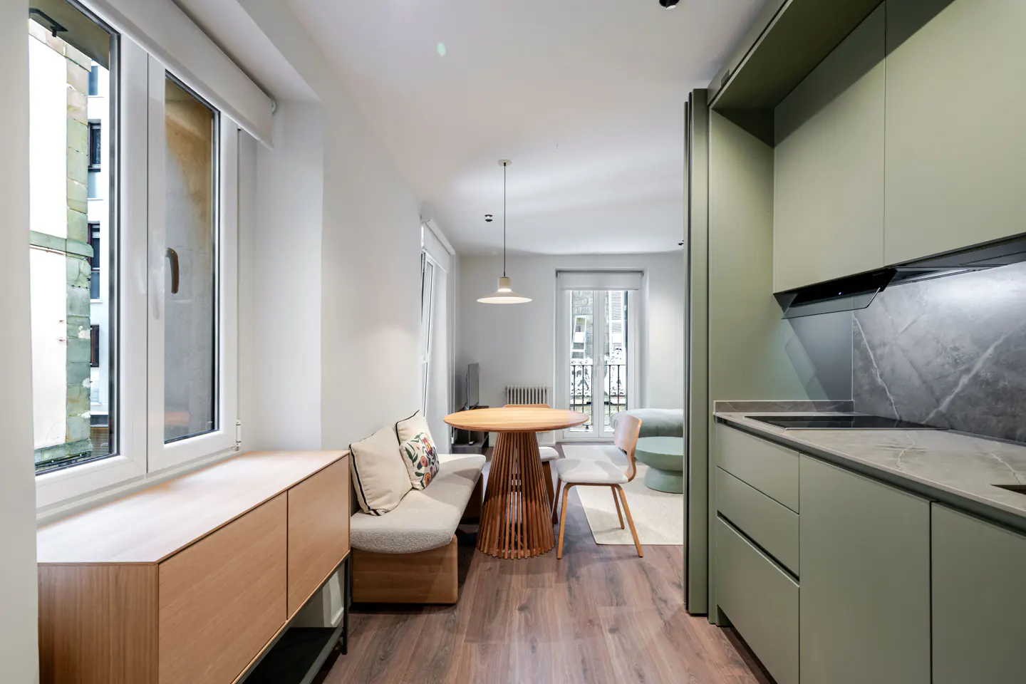 Interior view of a modern apartment with light green kitchen cabinets, a round wooden table, and a cushioned bench. Natural light streams through the windows.