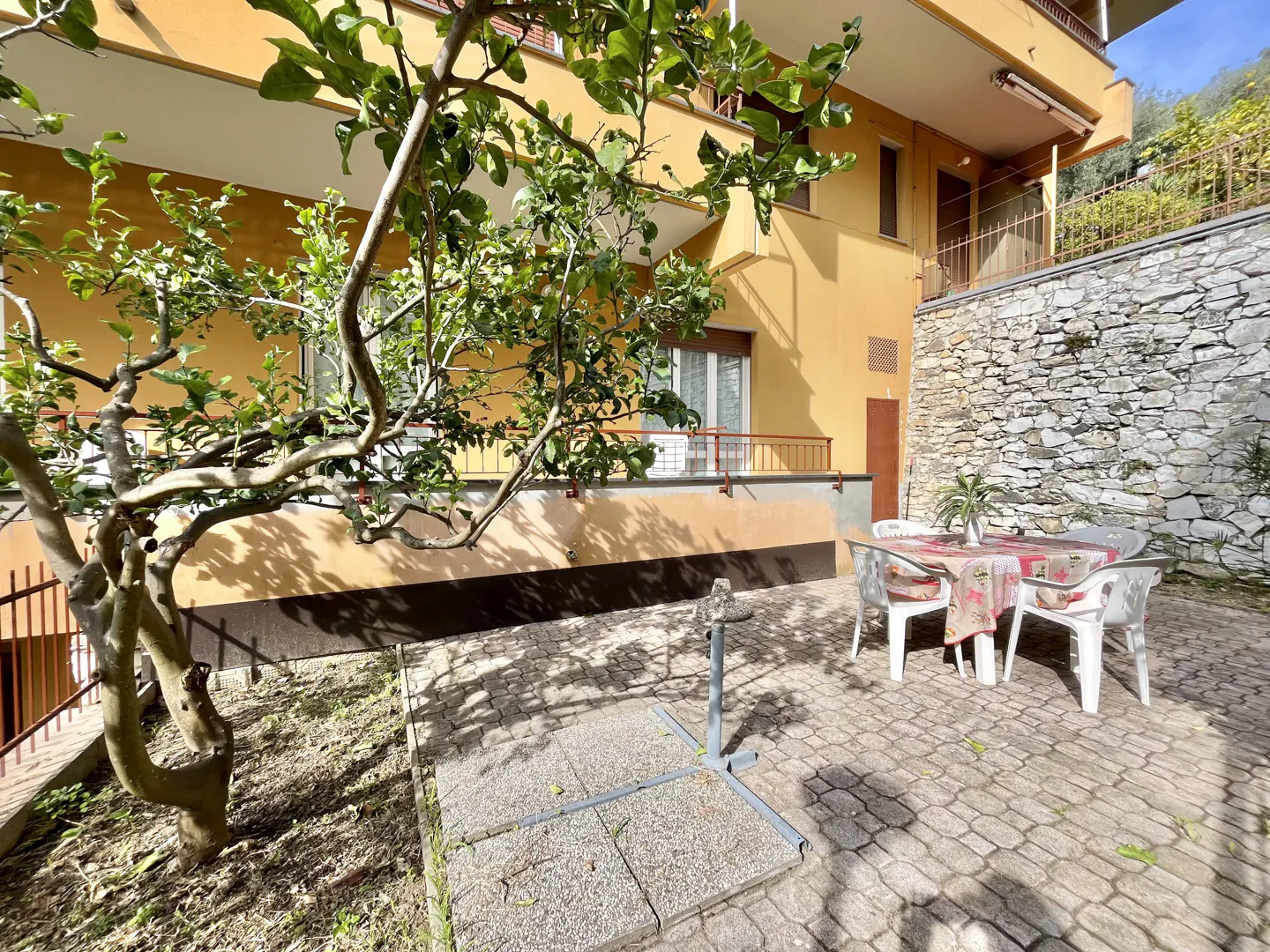 Outdoor patio with stone pavers, a table with a floral tablecloth, and a tree in front of a yellow building.