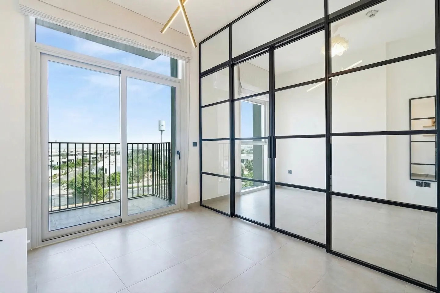 Bright room with a balcony view. Black framed glass doors divide the space. Outside, a black railing overlooks green trees and a blue sky.