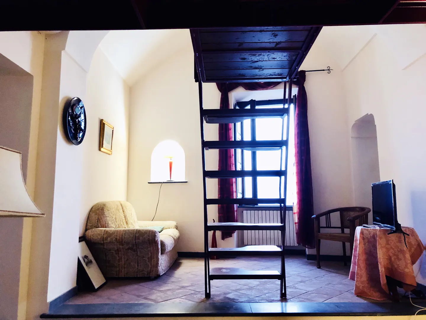 Cozy room with a loft bed accessed by a black ladder. A beige couch sits near a window niche with a candle. A TV and chair are on the right.