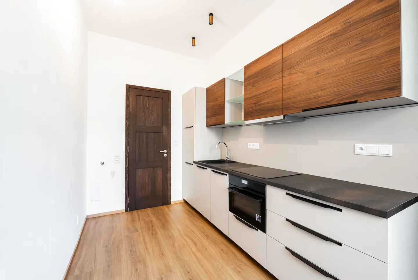 Modern kitchen with white lower cabinets, wood-grain upper cabinets, black countertops, and a dark wood door on wood floors.