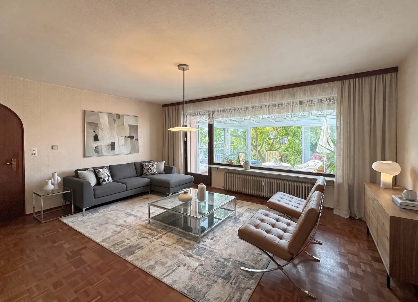 Modern living room with a grey sectional sofa, glass coffee table, two tan leather chairs, and a large window to a patio.