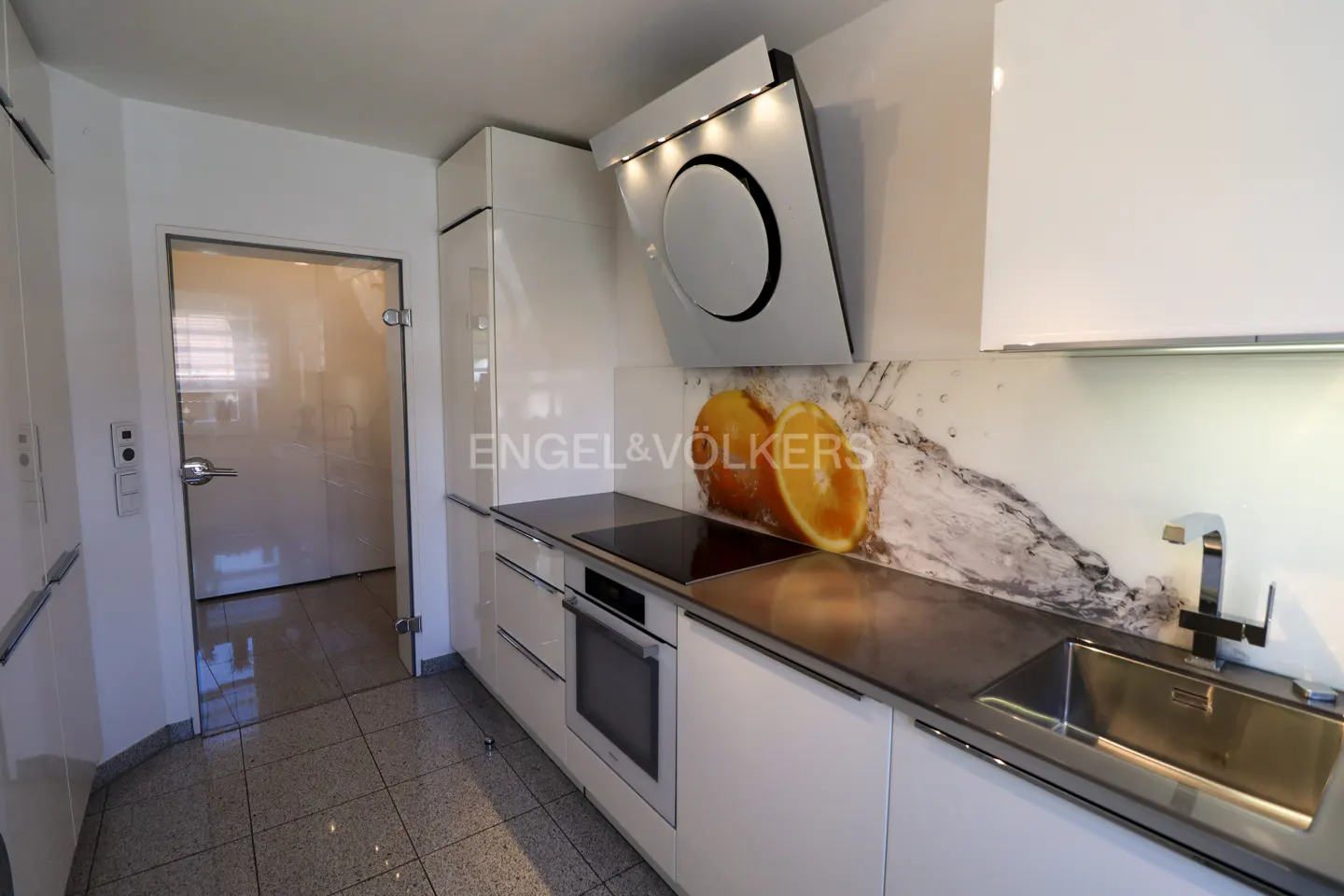 Modern white kitchen with stainless steel sink, black cooktop, and orange fruit backsplash. A glass door is visible on the left.