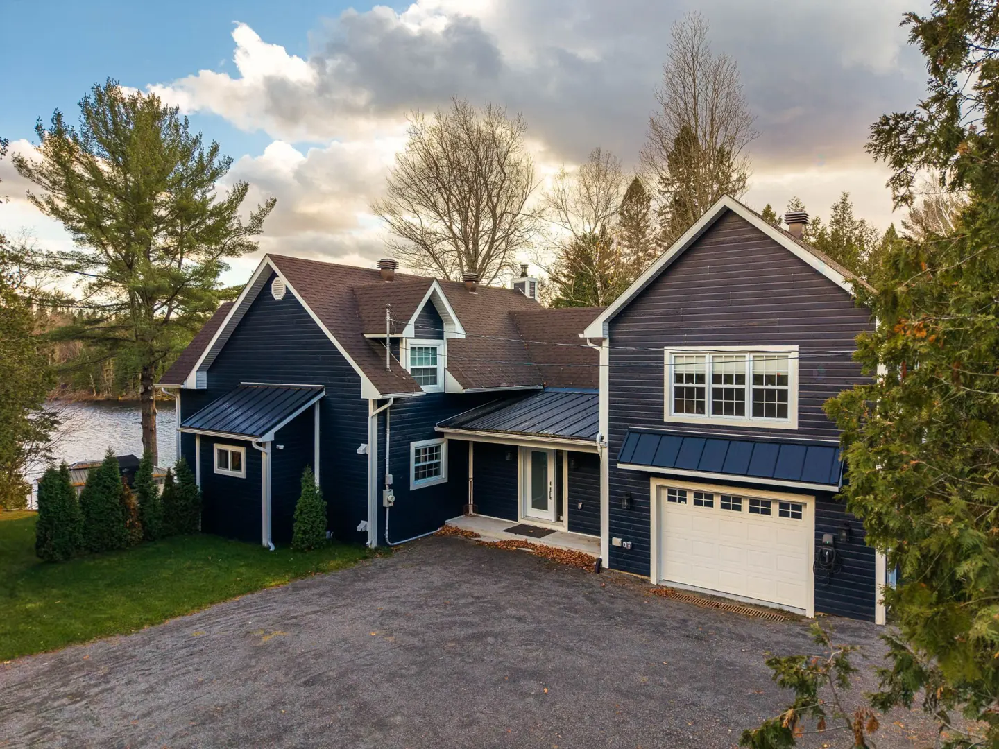 Two-story navy blue house with a brown roof and white garage door. A lake and trees are in the background.