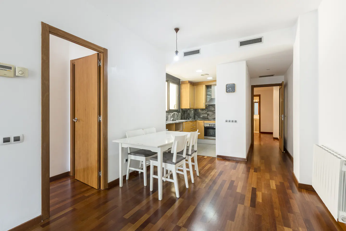 Bright apartment interior with hardwood floors, white walls, and a white dining table with four chairs. The kitchen is visible in the background.