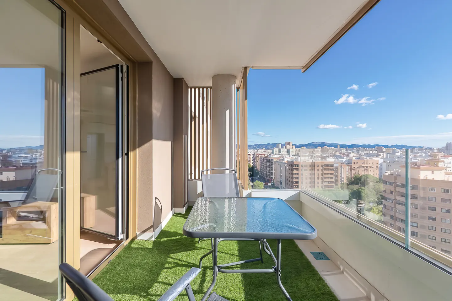 Balcony with artificial grass, table, and chairs overlooking a city skyline under a blue sky. Glass railing and sliding glass doors.