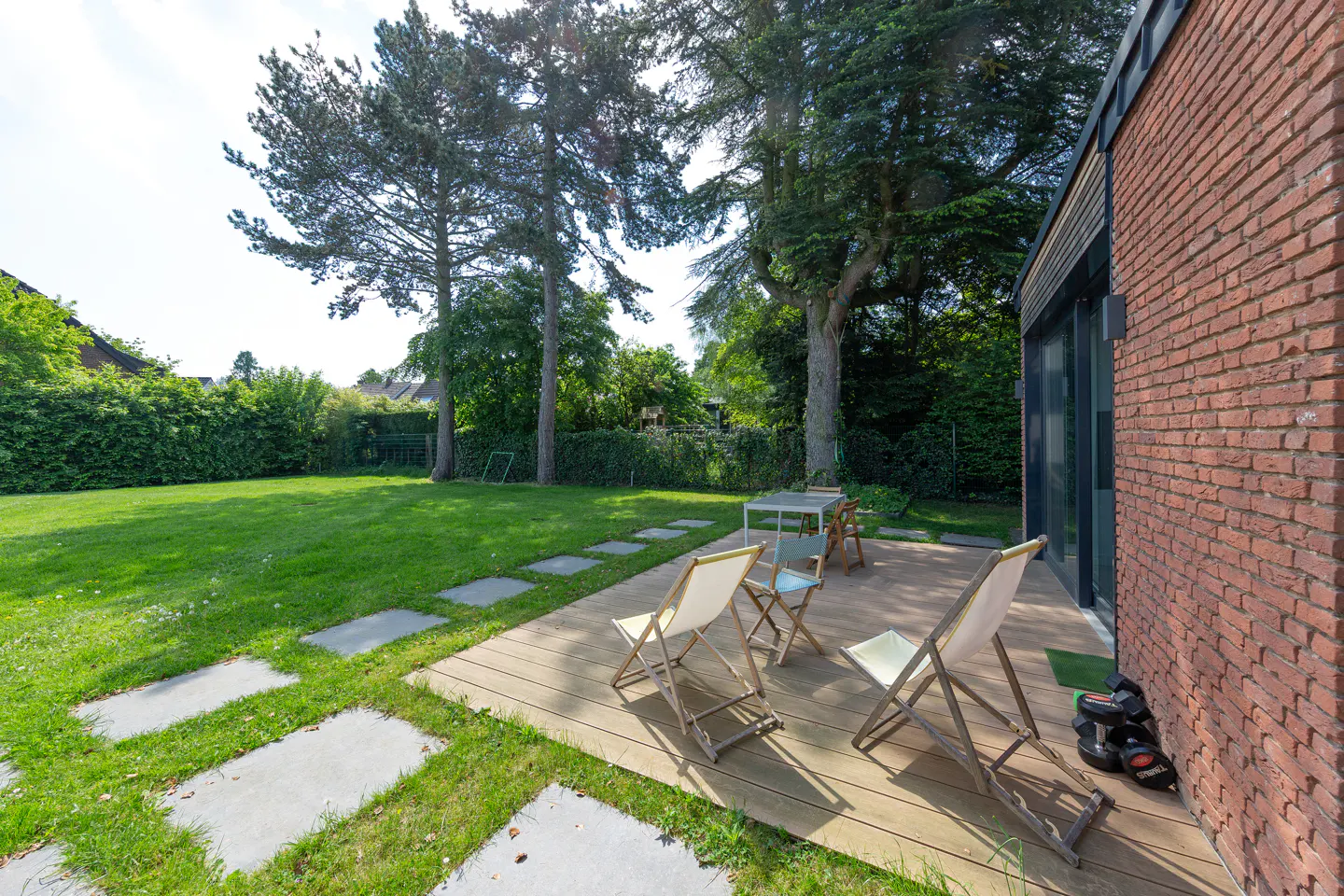 A wooden deck with lounge chairs and a small table next to a red brick house, overlooking a large, tree-lined green lawn.