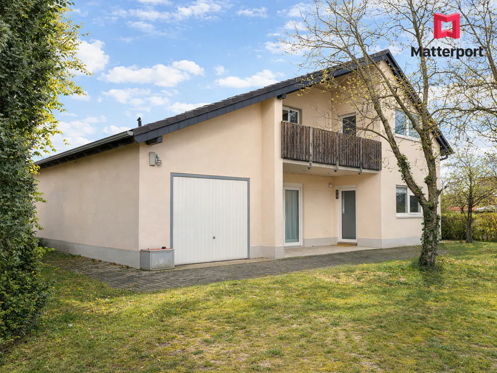 Two-story beige house with a garage, a balcony, and a green lawn on a sunny day.