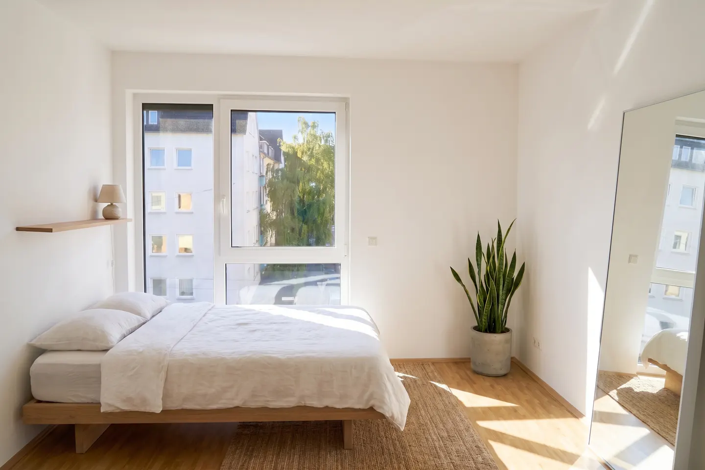 Minimalist bedroom with a wooden bed, white bedding, a potted snake plant, and a large floor mirror by a bright window.