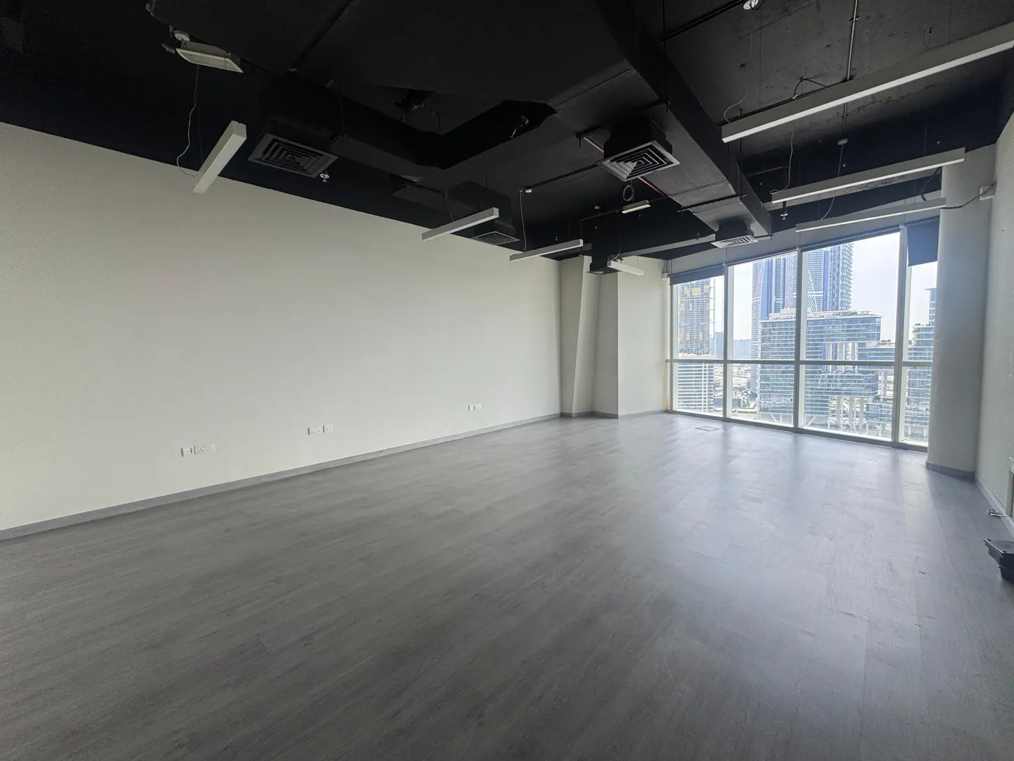 Empty office space with gray floors, white walls, and a black ceiling with lights. Large windows offer a city view.