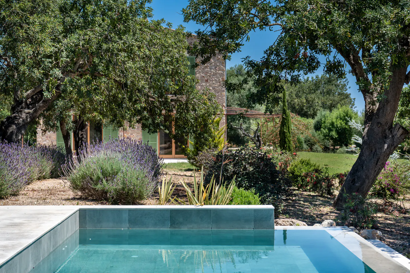 A turquoise pool reflects the sky, with a stone house, lavender bushes, and trees in the background.