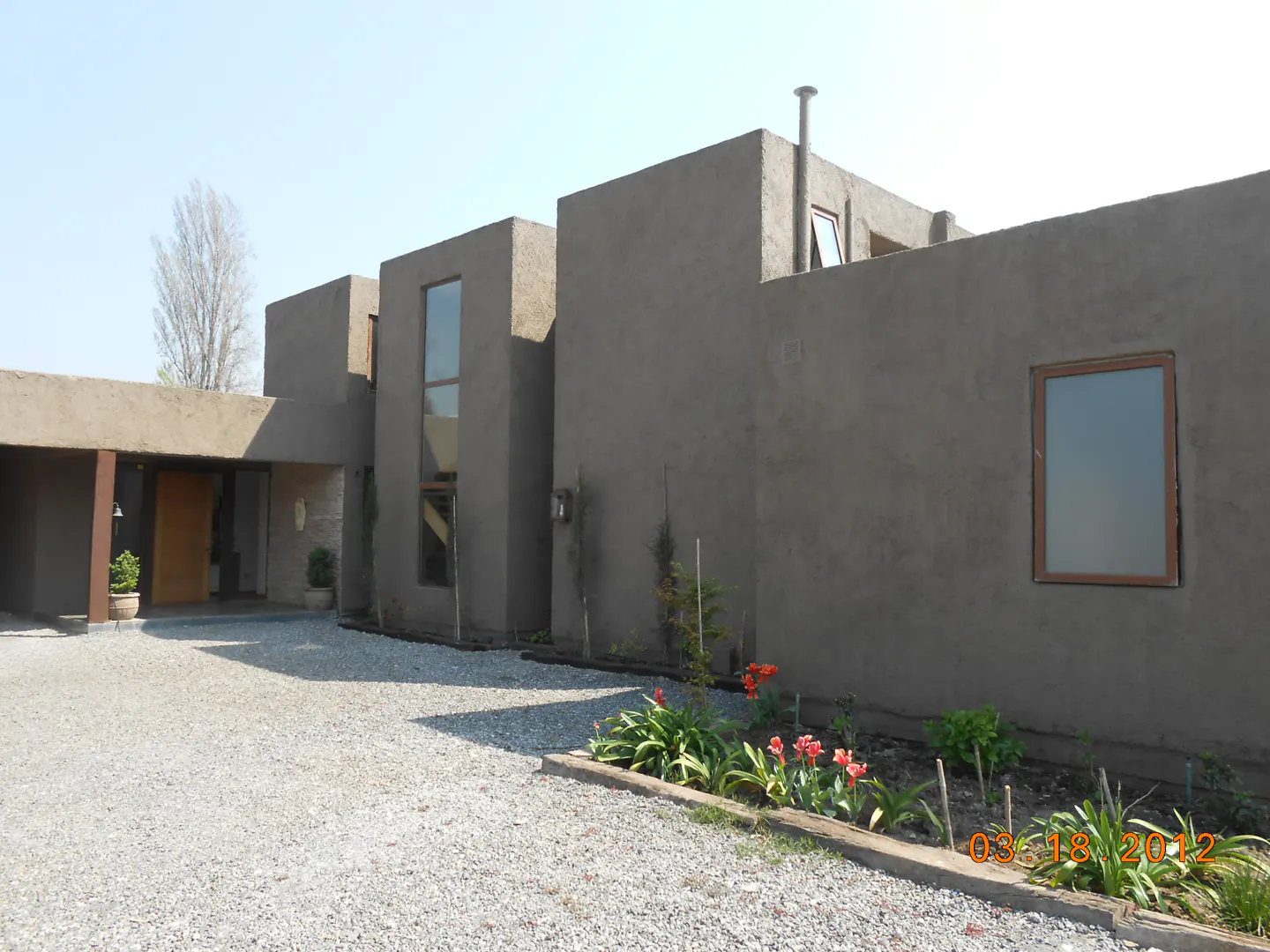 Modern gray house with flat roof, tall windows, and gravel driveway. A small flower bed with red tulips is in the foreground.