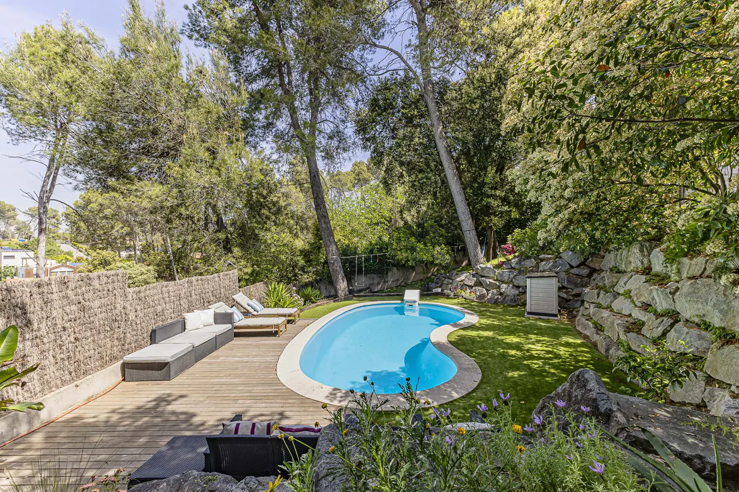 Outdoor pool area with a kidney-shaped pool, wooden deck, lounge chairs, and lush greenery. A stone wall and trees surround the space.