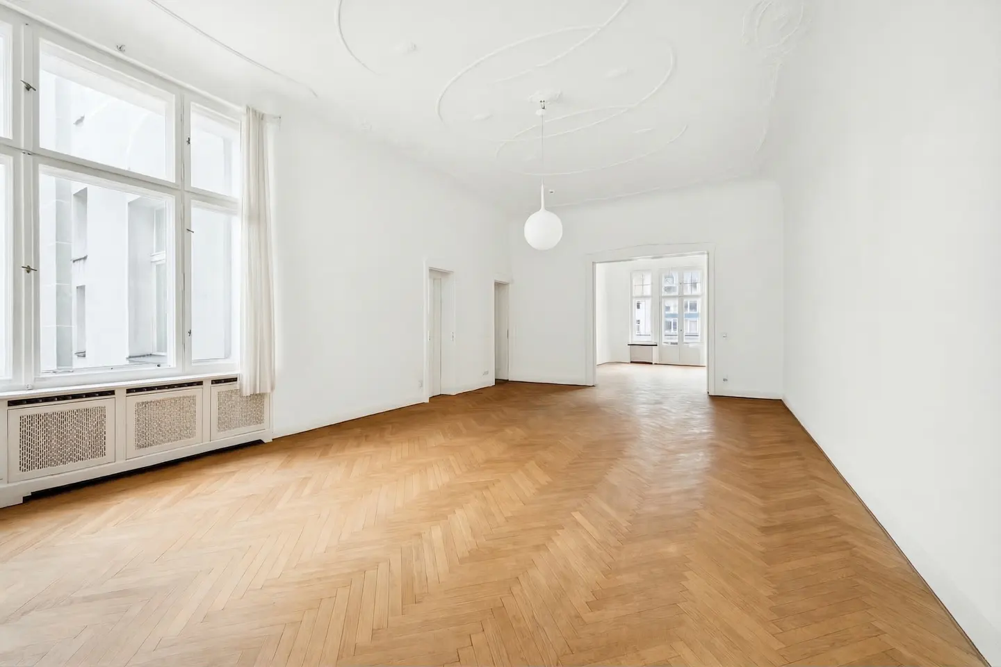 Bright, empty room with herringbone wood floors, white walls, and large windows. A white globe light fixture hangs from the ornate ceiling.