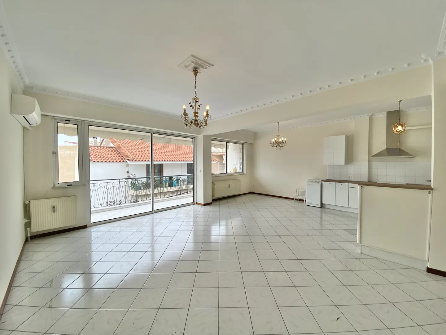 Bright, empty apartment with white tile floors, a balcony, and a small kitchen area with white cabinets. Chandeliers hang from the ceiling.