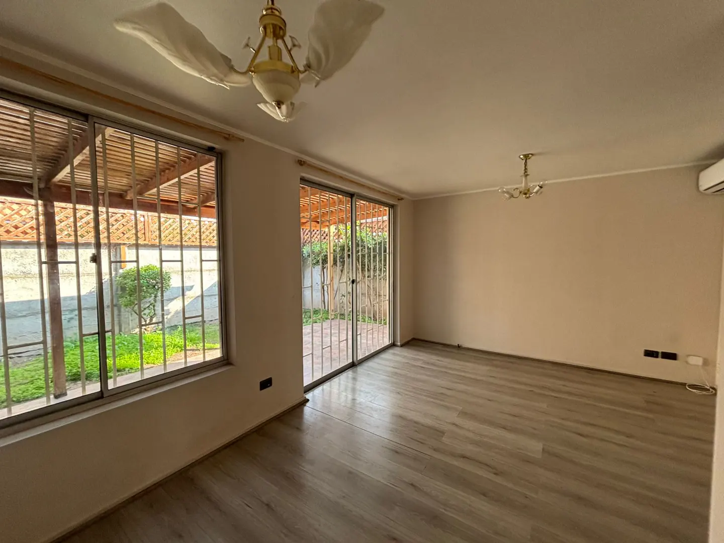 An empty room with light wood floors, beige walls, and a gold chandelier. Windows and sliding doors with bars look out onto a green yard.