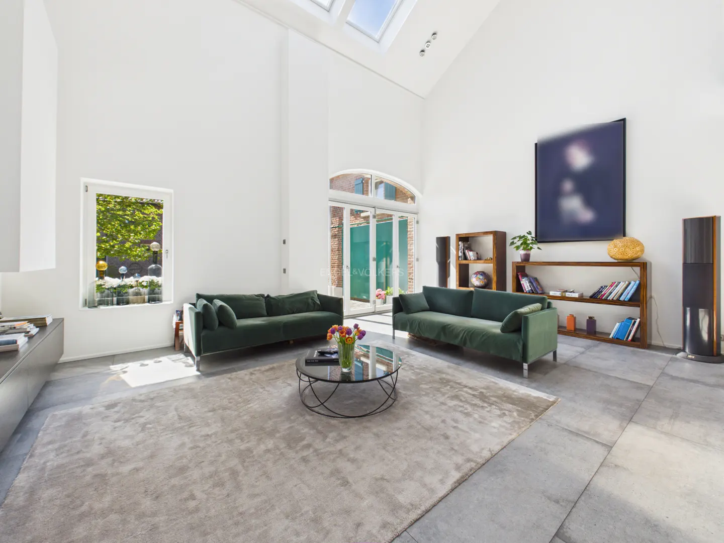 Bright living room with white walls, skylights, and concrete floors. Two green sofas face each other over a round glass table on a gray rug.