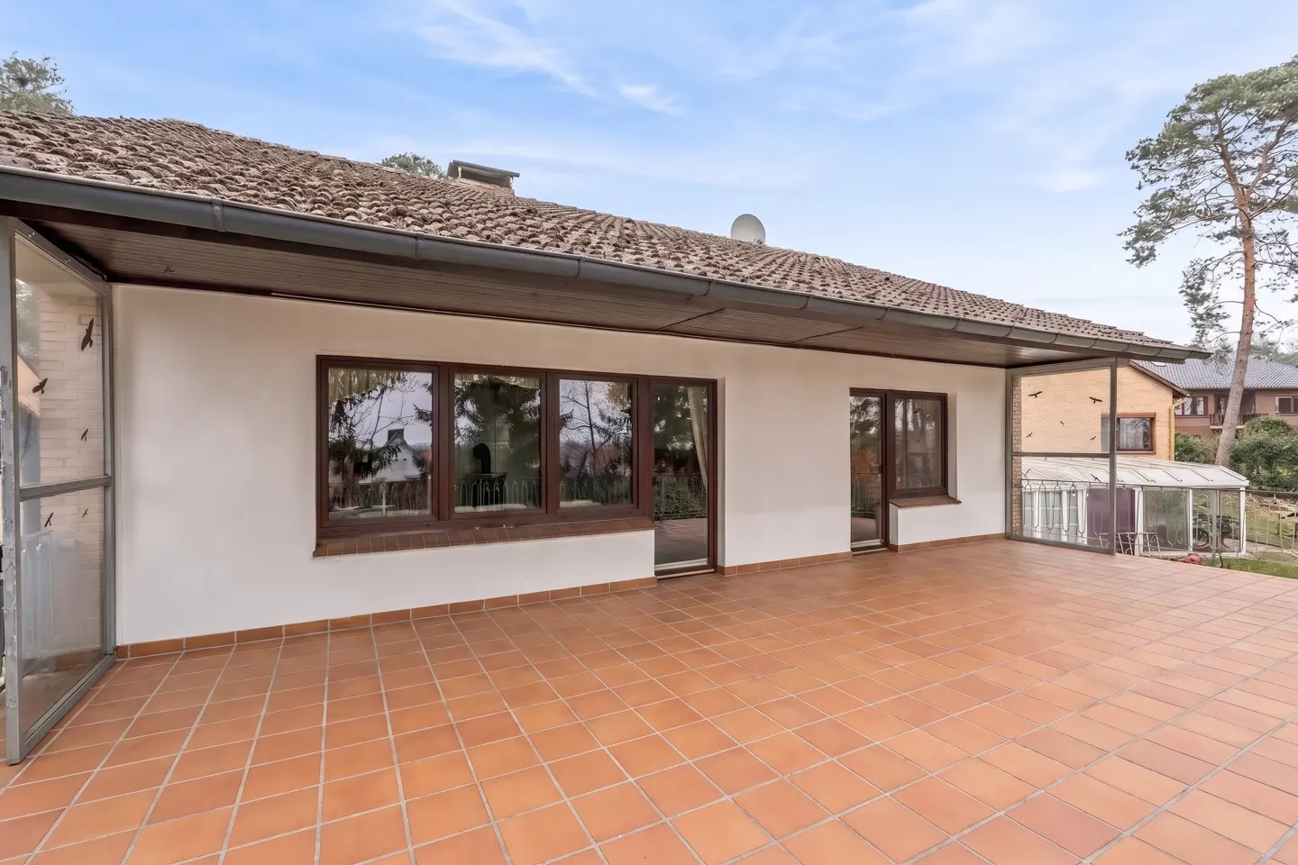 Exterior view of a house with a terracotta tiled patio, white walls, and dark-framed windows under a brown tiled roof.