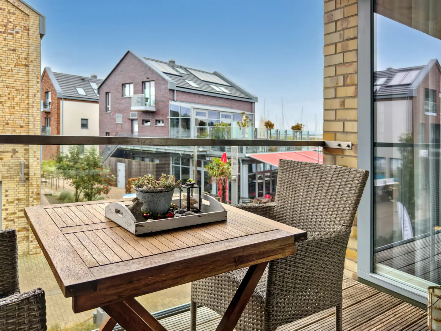 Balcony view with a wooden table, wicker chair, and potted plant. Buildings and blue sky are visible in the background.
