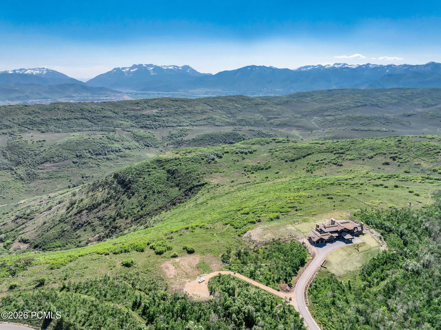 Aerial view of a large brown house on a green hillside with snow-capped mountains in the background under a blue sky.