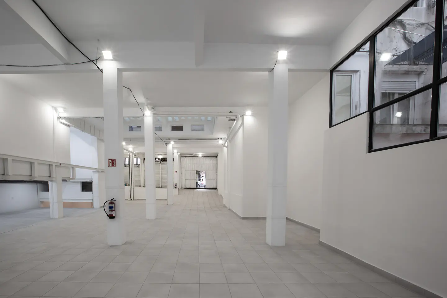 Bright, empty commercial space with white walls, gray tile floor, and support columns. A window with black frame is on the right.