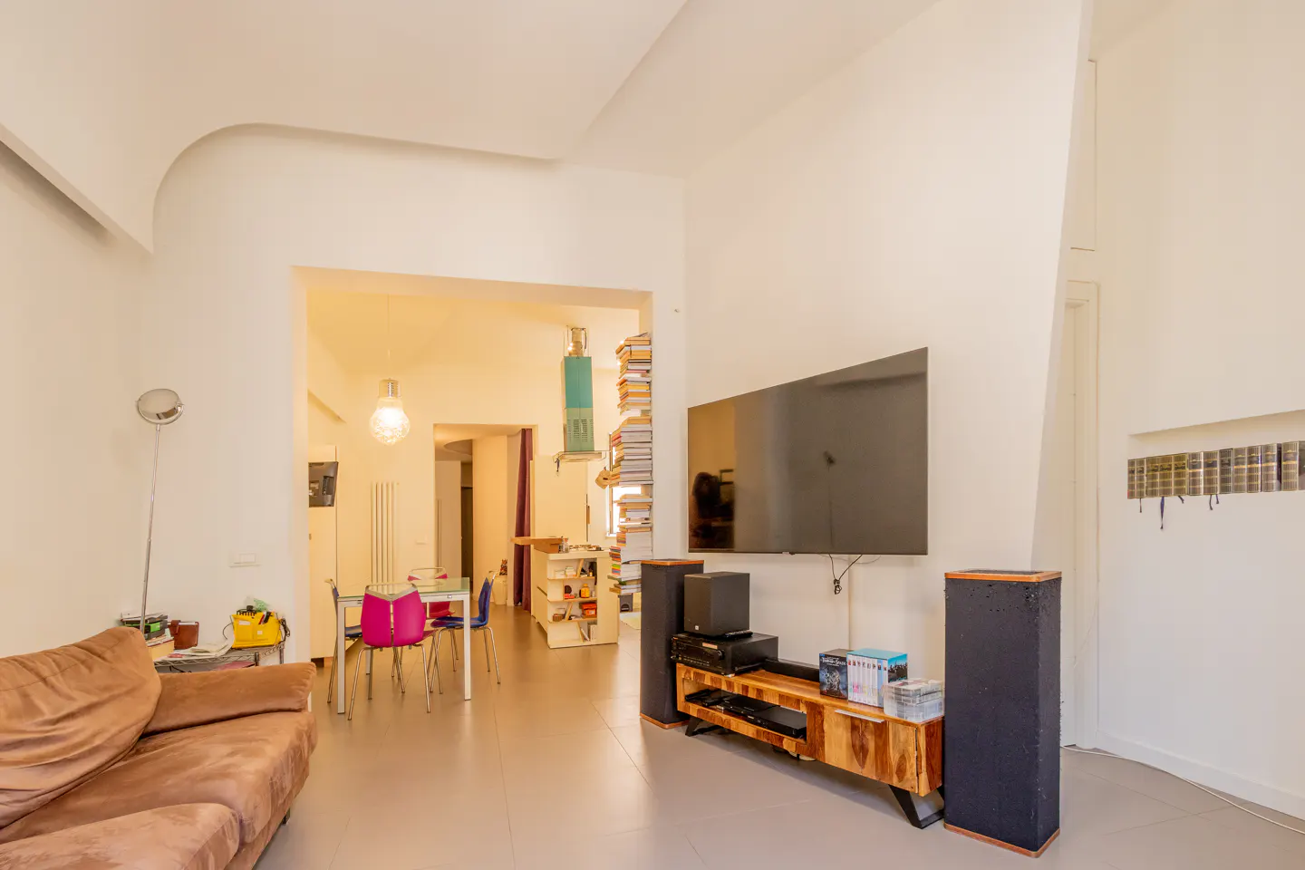 Modern living room with a brown sofa, wall-mounted TV, wooden media console, tall speakers, and a dining area in the back.