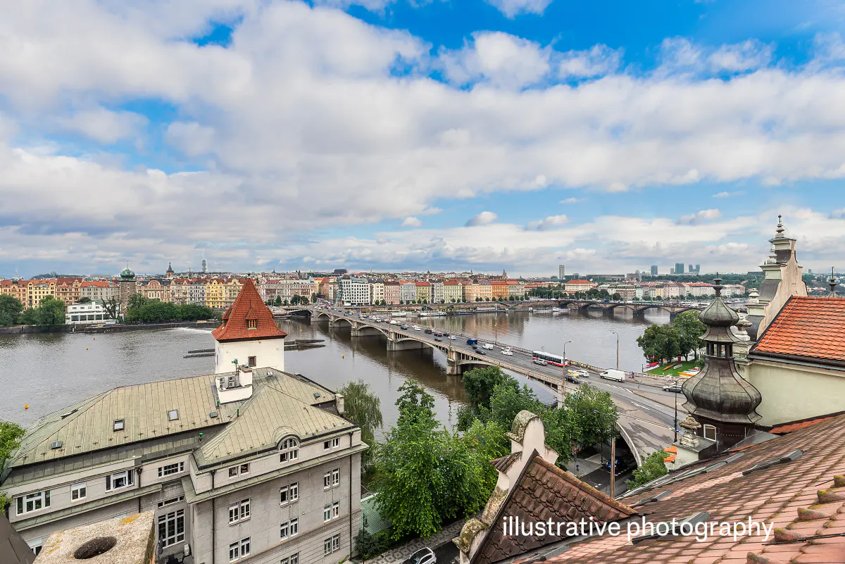 Cityscape view of Prague, Czech Republic, featuring a bridge over the Vltava River, buildings, and a cloudy blue sky.