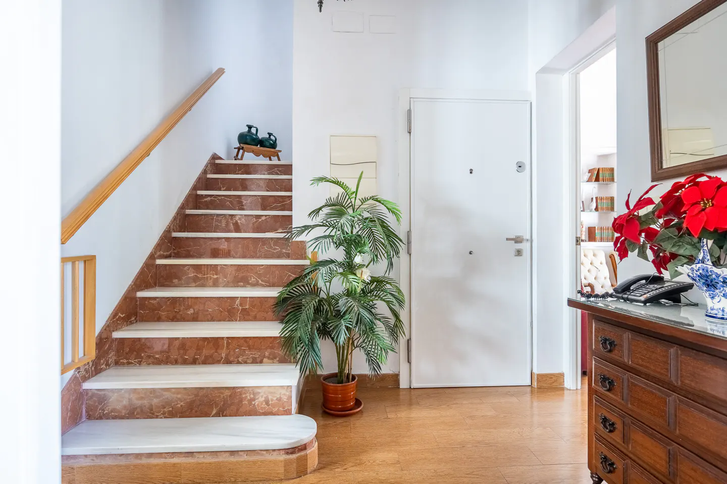 Bright entryway with red marble stairs, a white door, a potted palm, and a wooden chest with red poinsettias.