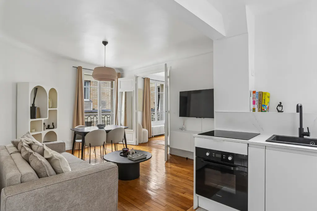 Bright open-plan apartment with a grey sofa, dining area, and modern white kitchen with black appliances and wood floors.