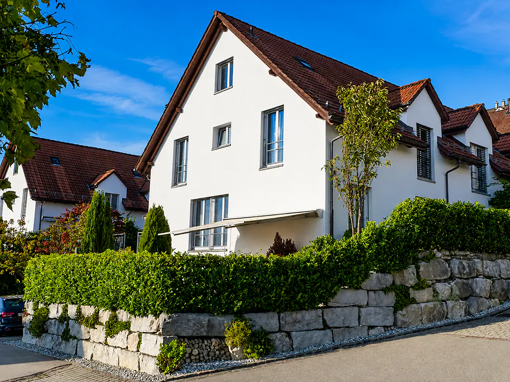 A two-story white house with a red tile roof, green hedge, and stone wall under a blue sky.