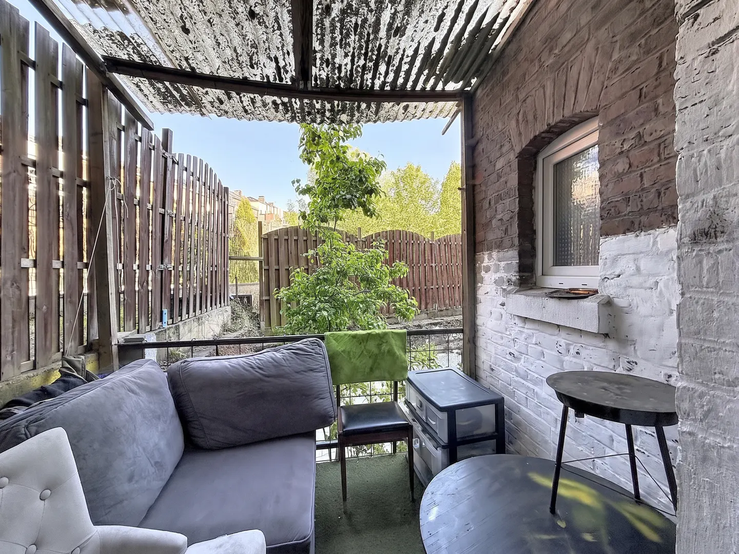 Covered porch with a gray couch, green chair, and black tables. A wooden fence and trees are visible in the background.