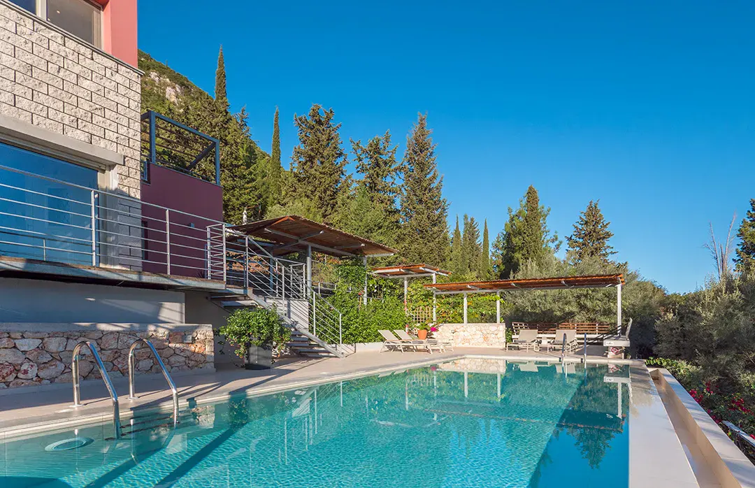 A modern villa with a stone facade and red accents features a swimming pool, patio seating, and lush trees under blue sky.