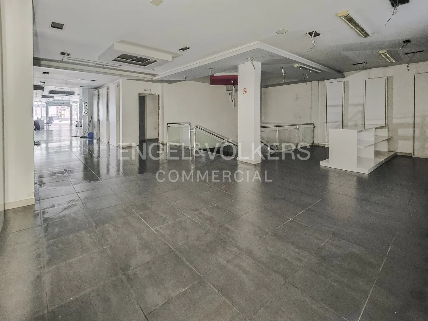 Empty commercial space with gray tile floor, white walls, and display shelves. Engel & Volkers logo in center.