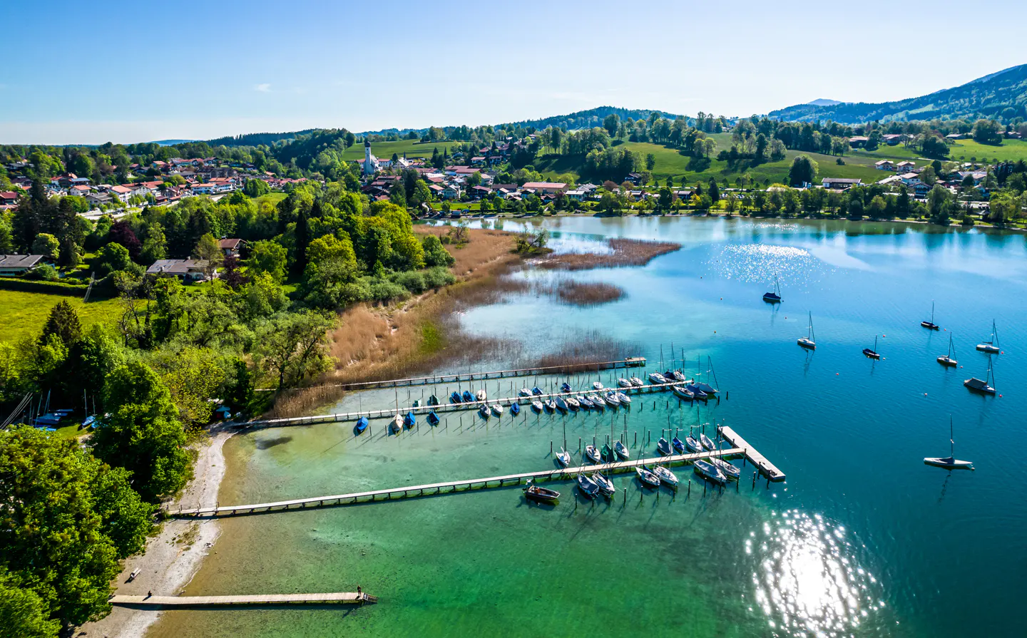 Aerial view of a lake with boats docked at a pier, surrounded by green trees and a town in the background.