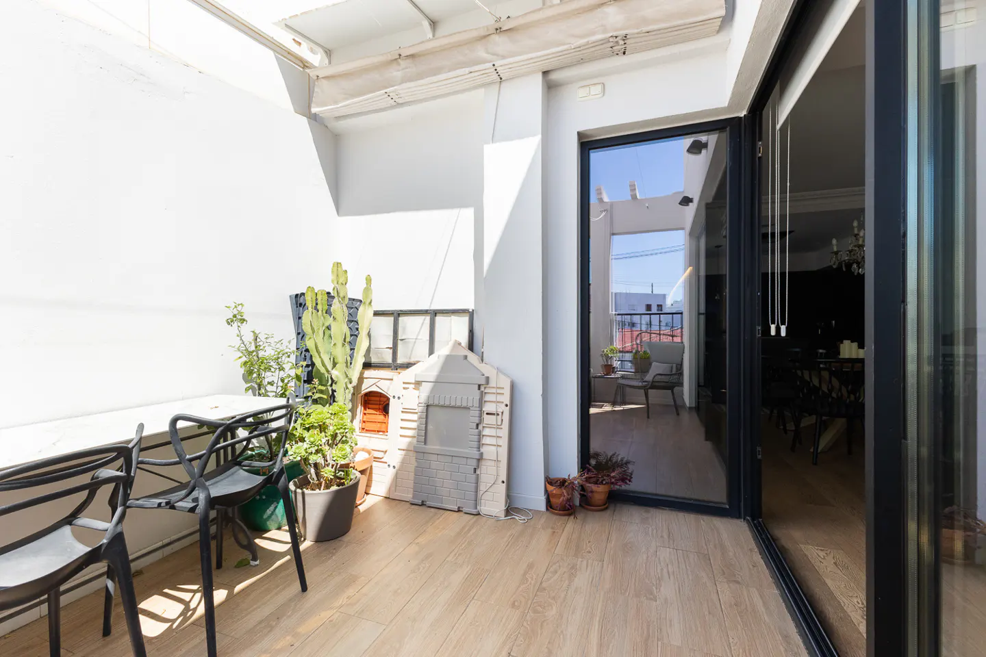 A bright balcony with black chairs, potted plants, and a child's playhouse. Sliding glass doors lead to an interior dining area.